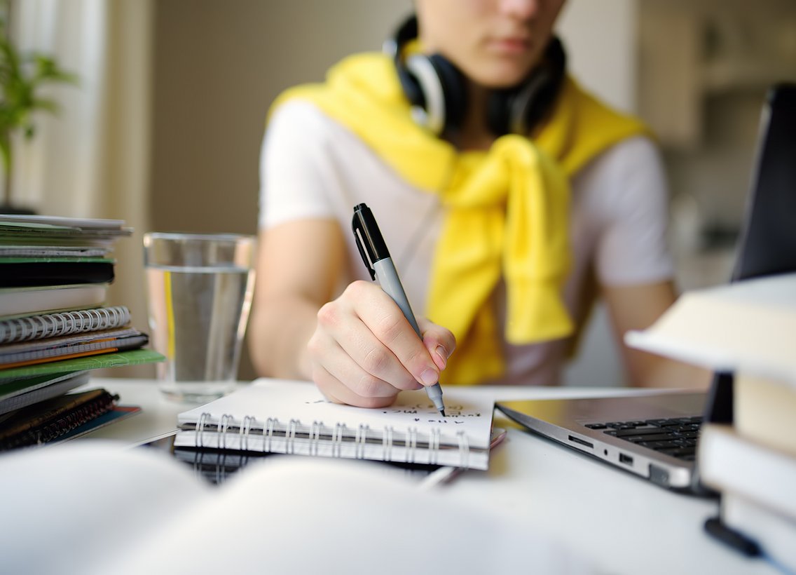 Im Hintergrund eine verschwommen sichtbare Person, im Vordergrund ein Schreibblock, ein Laptop und die Hand der Person, die einen Stift hält