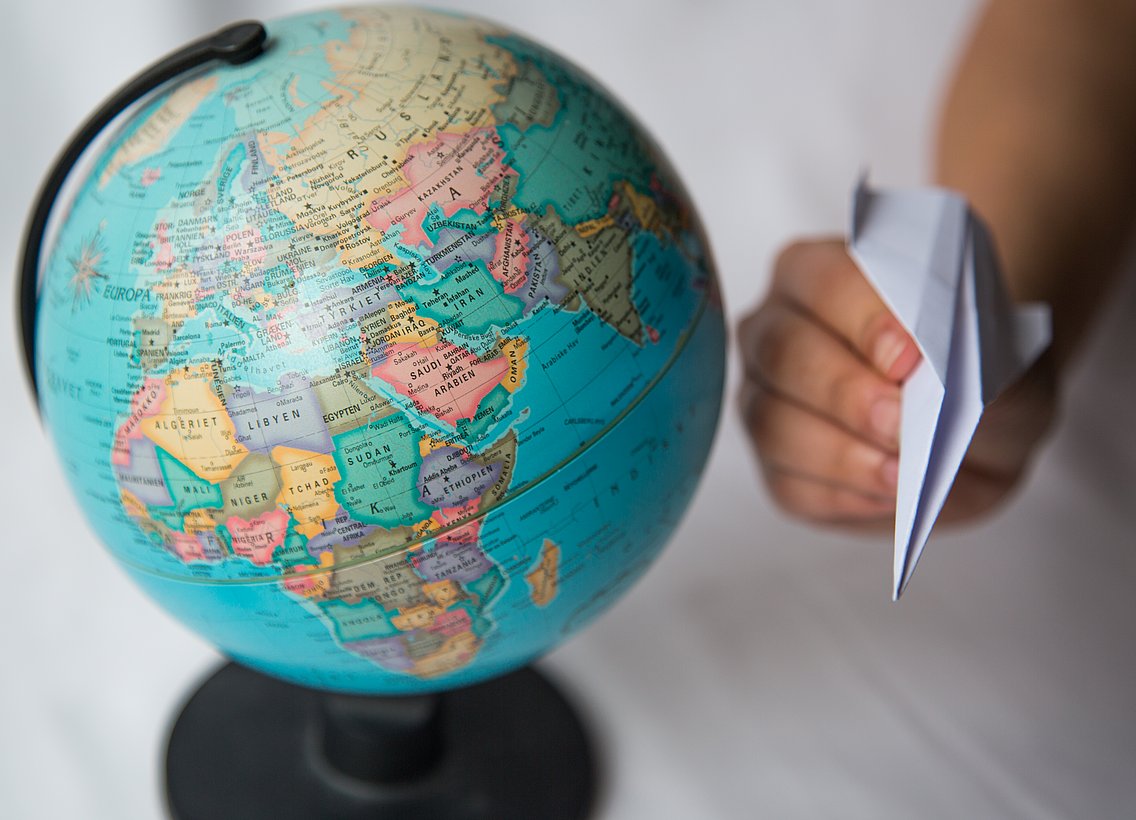 Ein Globus steht auf einem Tisch. Die Karte mit Europa ist im Vordergrund. Daneben ist eine Hand, die ein Papierflugzeug hält. 